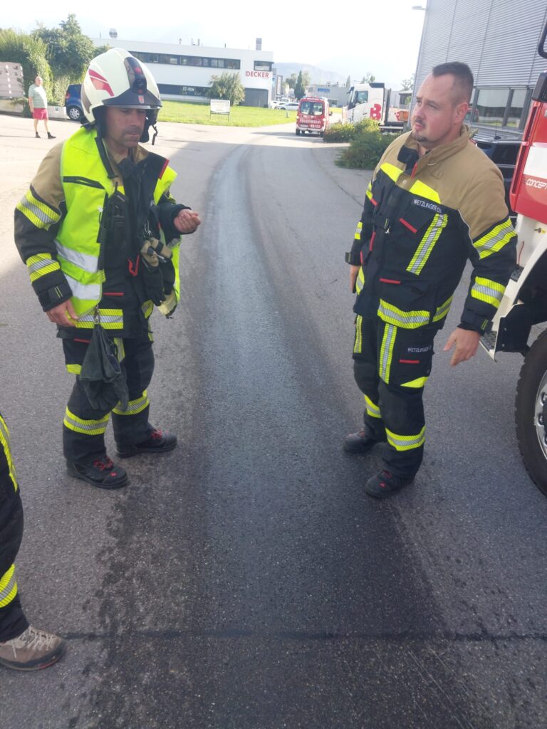 Mehrere Feuerwehrmänner, in Einsatzbekleidung, stehen rechts und link vor einer dunklen Flüssigkeits-Spur die sich in der Mitte einer Straße befinden